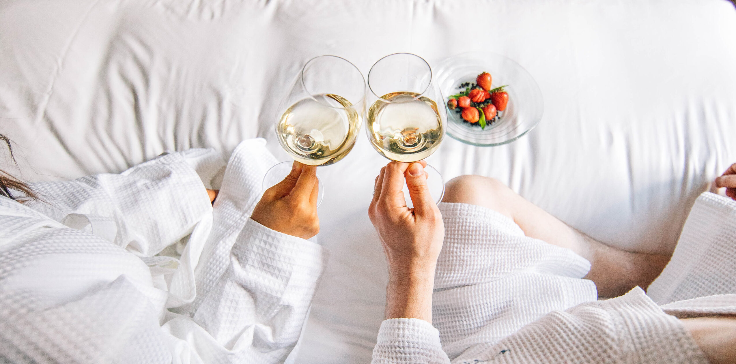 Couple in robes toasting wine on hotel bed